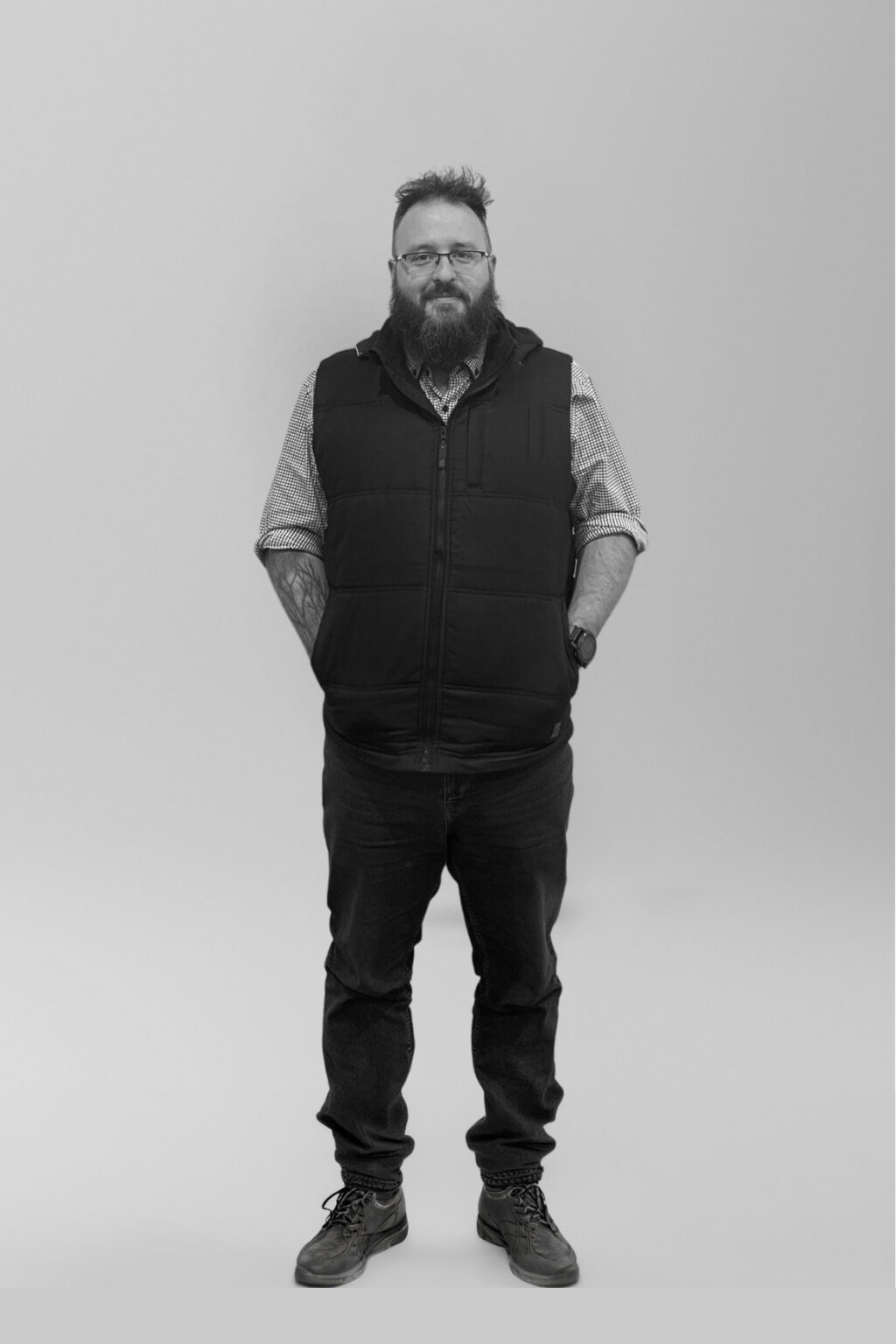 A bearded man wearing glasses, a checked shirt, a black waistcoat, dark jeans, and boots stands facing forwards with his hands behind his back against a plain grey background.