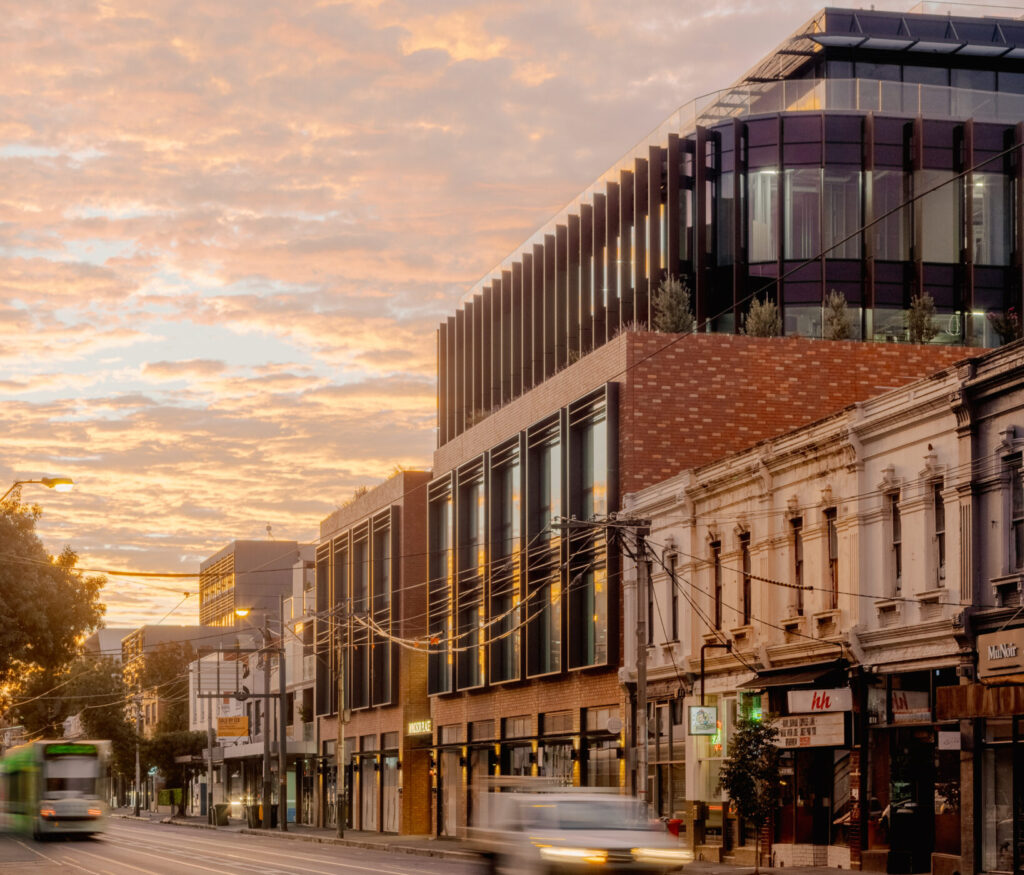 A city street at sunset with modern and historic buildings, blurred cars and a tram moving along the road, and a dramatic cloudy sky overhead.