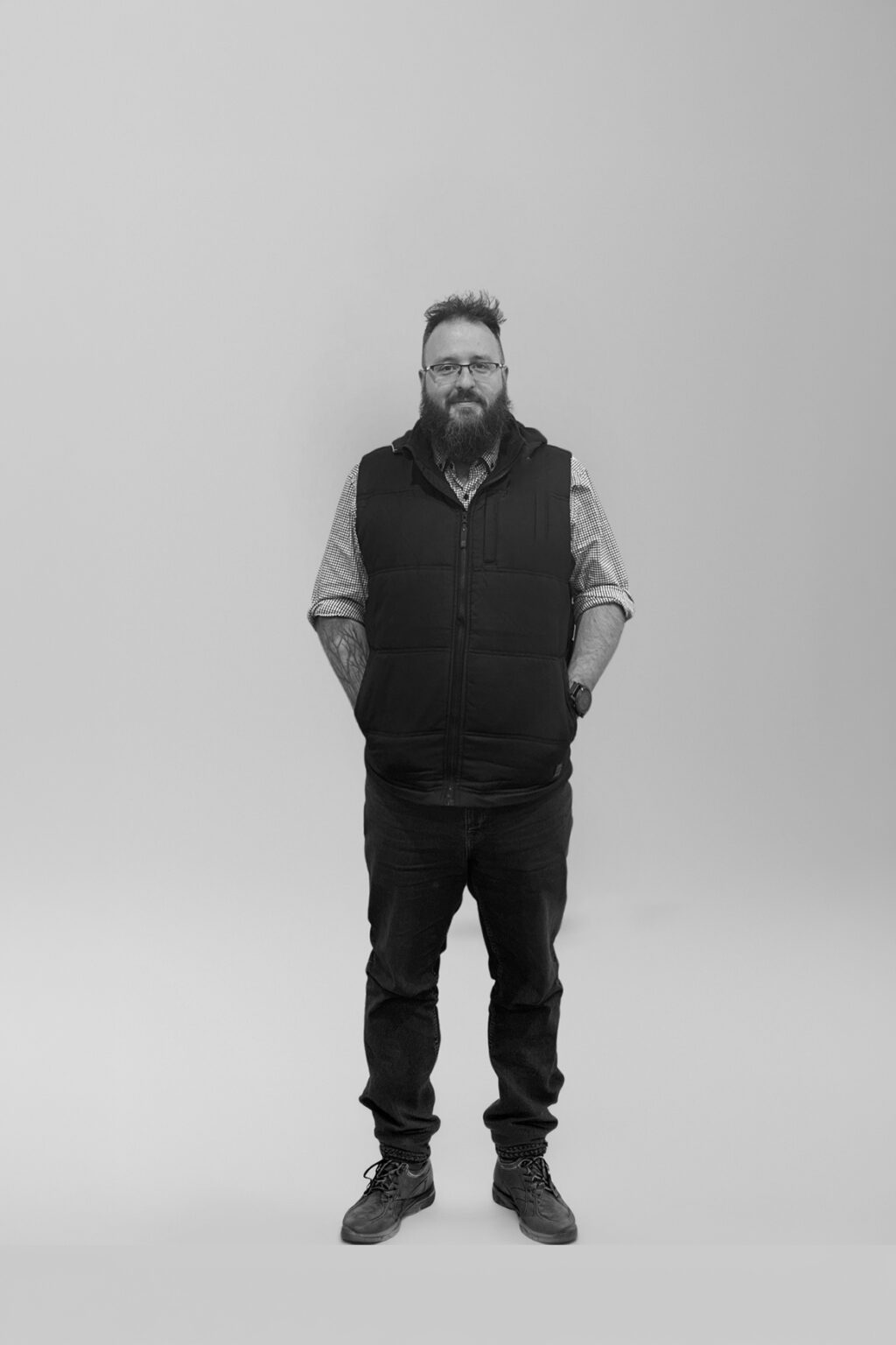 A bearded man wearing glasses, a checked shirt, a black waistcoat, jeans, and boots stands with his hands in his pockets against a plain light background.
