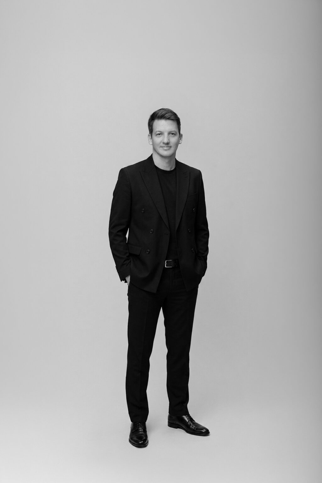 A person stands in front of a plain background, wearing a dark suit and dark shoes, with hands in their pockets. The image is in black and white.